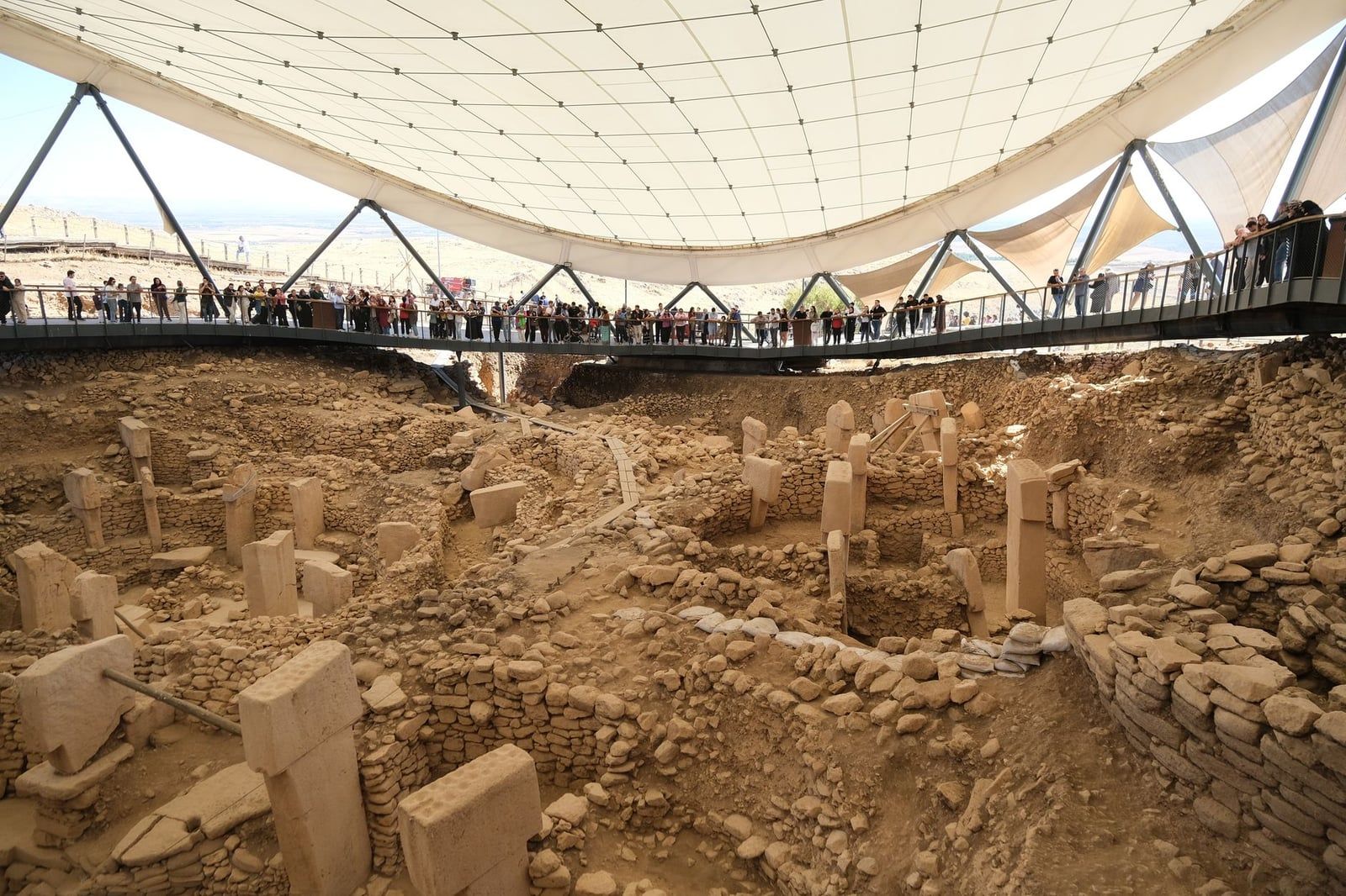Visitors stand along an elevated boardwalk beneath a wide protective canopy overlooking the ancient excavated stone enclosures of Göbekli Tepe.